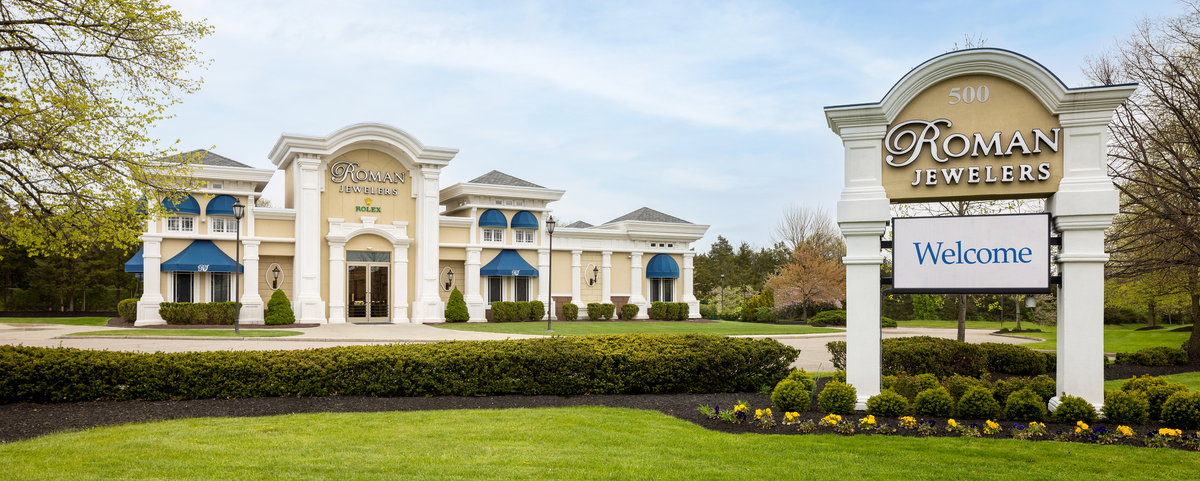 Roman Jewelers storefront with a large welcome sign in the foreground, set in a landscaped lawn with trees and blue sky.