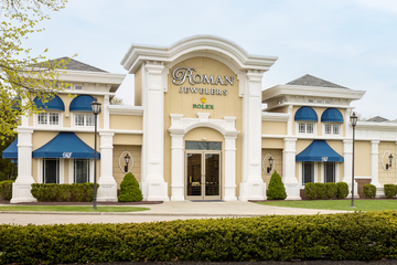 Front exterior view of the Roman Jewelers store, featuring a beige building with white columns, blue awnings, and a green lawn in the foreground.